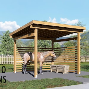 Puede incluir: Un refugio para caballos de madera con una silla de montar sobre un caballo, un abrevadero de madera y el texto "BAMBOO DESIGNS Home Plans & Designs". El refugio tiene un lado de listones y un techo sólido. El fondo incluye una valla blanca, árboles y una montaña.