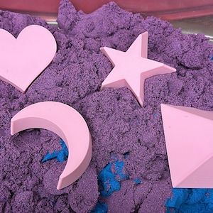 May include: Pink heart, star, crescent moon, and geometric shape toys in purple kinetic sand. The sand has blue accents. The toys are for sensory play and creative exploration.