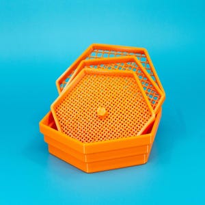 May include: Bright orange, hexagonal-shaped trays stacked on a blue background. The trays have a perforated design and a small, circular knob in the center. The trays appear to be made of plastic.