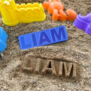 May include: A blue rectangular toy with the name "LIAM" printed on it, surrounded by colorful sandcastle molds in yellow, orange, and purple. The name "LIAM" is also imprinted in the sand.