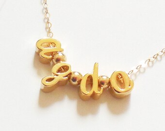 Könnte beinhalten: Eine goldfarbene Halskette mit dem Wort "geodo" in kursiven Buchstaben. Die Buchstaben sind durch kleine goldene Perlen verbunden. Die Halskette hat eine zarte goldene Kette.