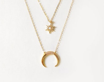 May include: Two-layered gold-toned necklaces. The top necklace features a starburst pendant with small, clear stones. The bottom necklace has a crescent moon pendant with a row of clear stones along the curve. Both necklaces have delicate chains.