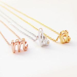 May include: Three delicate necklaces with initial charms. The charms are in gold, silver, and rose gold. The necklaces are layered and the charms are visible.
