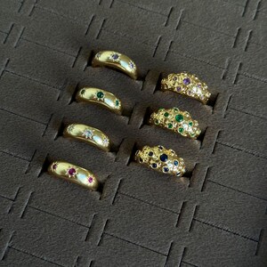 May include: A collection of gold rings displayed in a brown tray. The rings feature various gemstone embellishments, including rubies, emeralds, sapphires, and diamonds. Some rings have star-shaped designs.