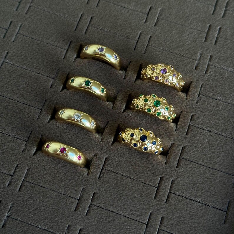 May include: A collection of gold rings displayed in a brown tray. The rings feature various gemstone embellishments, including rubies, emeralds, sapphires, and diamonds. Some rings have star-shaped designs.