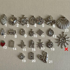 May include: A collection of silver-toned charms in various shapes, including hearts, suns, crosses, and a strawberry. Some charms feature intricate details and embellishments, perfect for jewelry making or crafting.