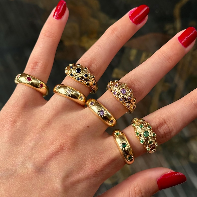 May include: A hand displays a collection of gold rings, each adorned with colorful gemstones. The rings feature various designs, including bands with single stones and ornate settings with multiple gems. The gemstones include red, blue, purple, and green, adding a vibrant touch to the jewelry.