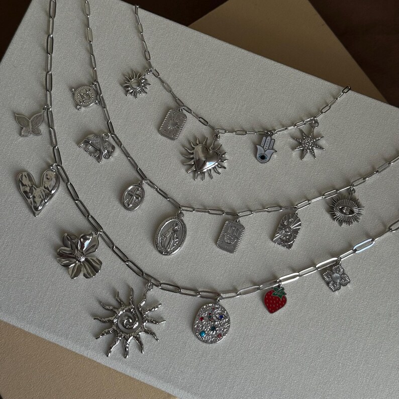 May include: Three silver charm necklaces with various pendants, including a butterfly, heart, flower, sun, and strawberry. The charms are detailed and the necklaces have a delicate chain design. The necklaces are displayed on a neutral surface.