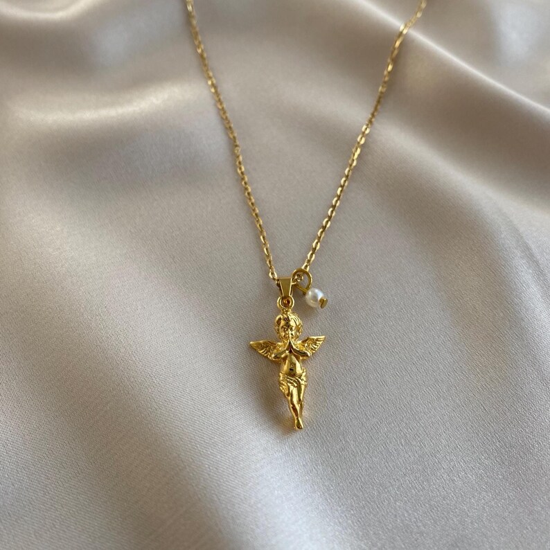 May include: A gold necklace with a cherub pendant and a small pearl. The cherub has outstretched wings. The chain is fine, and the pendant is detailed. The necklace is shown against a soft, off-white background.