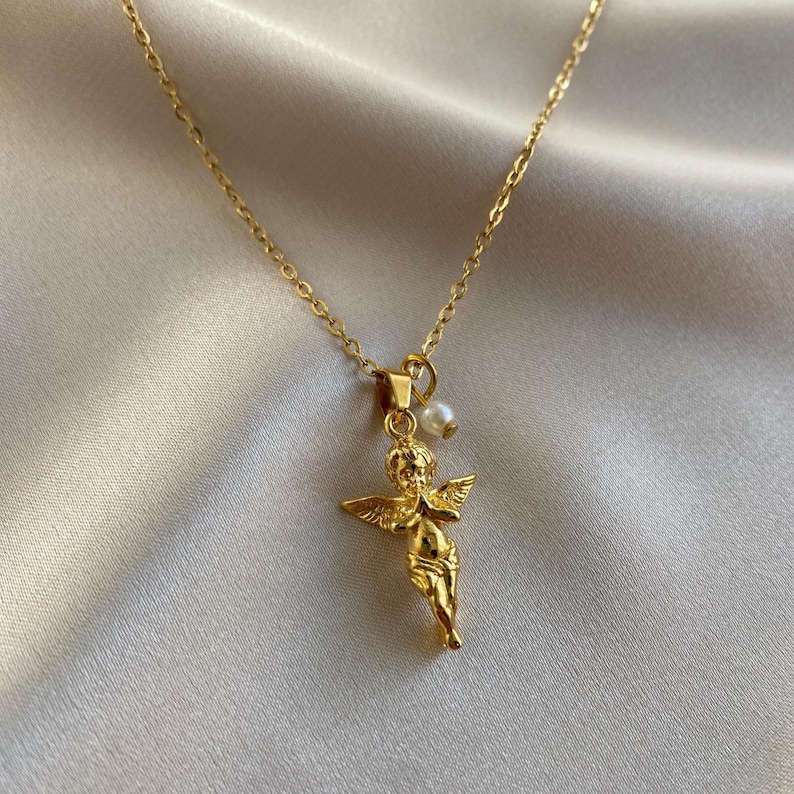 May include: A gold-coloured necklace featuring a cherub pendant with outstretched wings and a small pearl. The delicate chain and pendant are made of a shiny, metallic material. The cherub is in a standing pose.