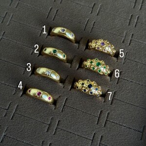 May include: A collection of seven gold rings, each with a unique design. Some rings feature gemstones like emeralds, rubies, sapphires, and diamonds. The rings are displayed in individual slots within a dark brown case.