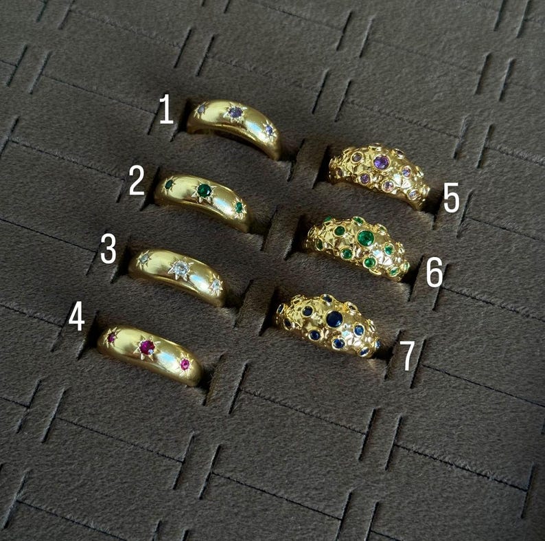 May include: A collection of seven gold rings, each with a unique design. Some rings feature gemstones like emeralds, rubies, sapphires, and diamonds. The rings are displayed in individual slots within a dark brown case.