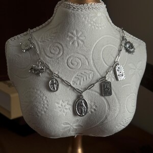 May include: A silver-toned charm necklace with various pendants, including a turtle, elephant, cross, and a religious figure. The necklace is displayed on a white mannequin bust, showcasing its design and details. The chain has a link style.