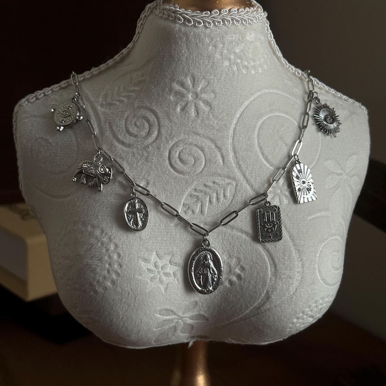May include: A silver-toned charm necklace with various pendants, including a turtle, elephant, cross, and a religious figure. The necklace is displayed on a white mannequin bust, showcasing its design and details. The chain has a link style.
