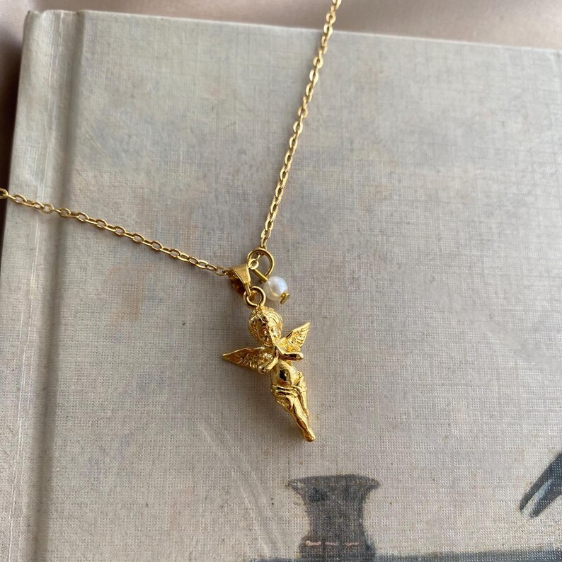 May include: A gold-coloured necklace with a small cherub pendant and a pearl. The pendant is detailed with outstretched wings. The fine chain is displayed on a vintage book. The necklace is a delicate piece of jewellery.