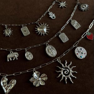 May include: Silver charm necklaces with various pendants, including a sun, heart, flower, and elephant. The charms are attached to a chain. The background is a solid brown color.