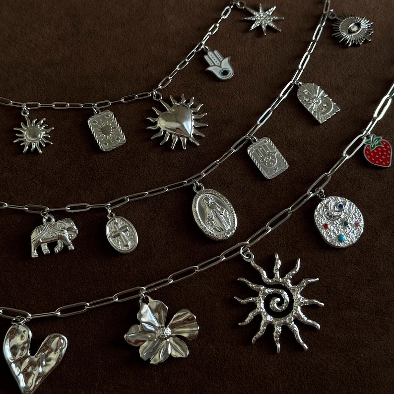 May include: Silver charm necklaces with various pendants, including a sun, heart, flower, and elephant. The charms are attached to a chain. The background is a solid brown color.