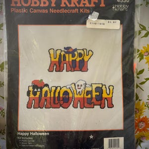 May include: A sealed Hobby Kraft plastic canvas needlecraft kit. The kit features the words "HAPPY HALLOWEEN" in a cross-stitch design. The text is in yellow, orange, and blue. The kit includes plastic canvas and acrylic yarn.
