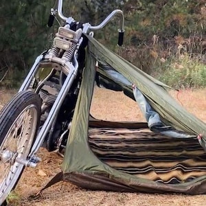 May include: A vintage motorcycle with a chrome frame and a black tire is parked next to a small olive green tent. The tent has a striped interior and is set up on dry grass, suggesting an outdoor camping scene.