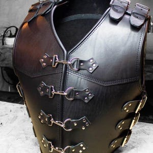 Puede incluir: Chaleco de cuero negro con múltiples correas y hebillas. El chaleco presenta un diseño de cuello en V y se asegura con herrajes de metal plateado. El cuero parece liso y el chaleco tiene una silueta estructurada.