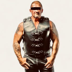 May include: Black leather vest with decorative metal clasps. The vest has a V-neck design and is sleeveless. The vest is worn over a black shirt and black pants. The vest is a classic biker style.