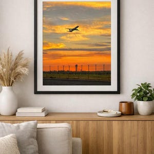 May include: A framed wall art print featuring a silhouette of an aeroplane taking off against a vibrant sunset sky with orange and yellow clouds, above a dark runway and a perimeter fence with a distant control tower visible on the horizon.