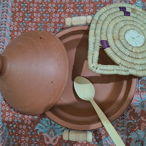 May include: A terracotta tagine pot with a matching lid and a woven lid, alongside a wooden spoon. The tagine is a traditional North African cooking vessel. The woven lid has purple accents.