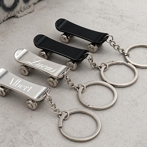 Könnte beinhalten: Vier Skateboard-Schlüsselanhänger in einer Reihe. Zwei sind aus poliertem silberfarbenem Metall mit den eingravierten Namen Albert und Lucas auf dem Deck, zwei sind einfarbig schwarz. Jeder hat eine Metallkette und einen Schlüsselring.