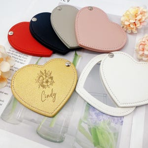 以下が含まれることがあります： 赤、ネイビー、グレー、ピンク、ゴールド、白の様々な色のハート型のコンパクトミラーのコレクション。 金色のミラーには花の模様と「Cindy」という名前が刻まれています。 各ミラーには、吊り下げ用の小さな金属ループが付いています。