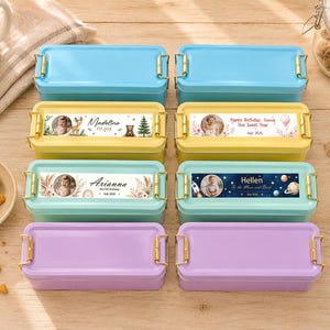 May include: An overhead shot of eight colorful lunchboxes with gold-colored clasps. The lunchboxes are blue, yellow, mint green, and purple. Some lunchboxes have personalized labels with a photo and text. A mug and cookies are in the background.