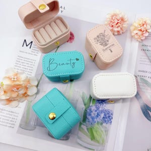 May include: Several small jewellery boxes in pastel colours. The boxes are pink, blue, and white, with gold clasps. One box is open, showing ring slots. The word "Beauty" is written on one box. Another box has the name "Sammie".