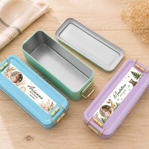 Op de afbeelding: Drie rechthoekige metalen lunchboxen in blauw, groen en paars, met goudkleurige sluitingen. De blauwe en paarse dozen hebben gepersonaliseerde labels met een foto en naam. De groene doos is open en toont een zilveren interieur.