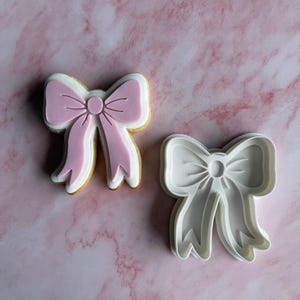 May include: A bow-shaped biscuit and a matching cookie cutter. The biscuit has a pink bow design with white and yellow accents. The cookie cutter is white plastic and has the same bow shape. The background is a pink marble pattern.