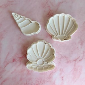 May include: Three white seashell-shaped cookie cutters. One is a conch shell, and the other two are scallop shells. The cutters have a simple design with a thin gold outline. The cutters are made of plastic and are on a pink marble surface.