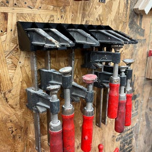 May include: A collection of woodworking clamps with red handles and black metal components, mounted on a wooden wall. The clamps are of various sizes and styles, designed for holding materials together during woodworking projects.