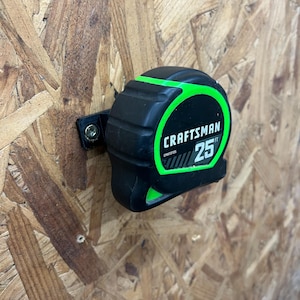 May include: A black and green Craftsman 25 ft tape measure mounted on a wooden surface. The tape measure has a green border and the number 25 in green. The Craftsman logo is visible.