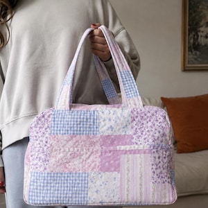 Op de afbeelding: Een gewatteerde draagtas met een patchworkontwerp in tinten lichtpaars, lavendel en wit. Het stofpatroon bevat kleine bloemenprints, gingham-ruitjes en verticale strepen. De tas heeft twee bijpassende stoffen handvatten.