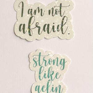 May include: Two stickers with inspirational quotes. The top sticker reads "I am not afraid" in a handwritten font. The bottom sticker says "strong like aelin" in a teal script. Both stickers have a holographic, star-like background.