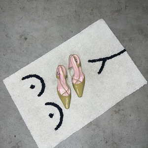 May include: A pair of olive green pointed-toe slingback heels with thin crisscross straps and light pink insoles, resting on a rectangular off-white tufted rug featuring a black abstract line drawing of a face with two eyes and a mouth.
