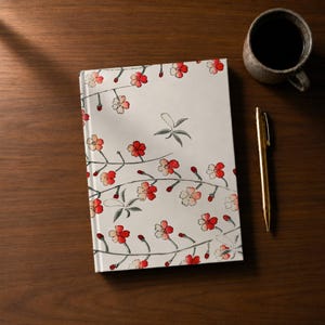 May include: A white journal with a floral design featuring red and white blossoms and green leaves. A gold pen and a mug of coffee sit beside the journal on a dark wooden surface. The journal's cover is decorated with a delicate, repeating pattern.