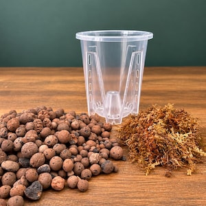 Può includere: Un vaso per piante in plastica trasparente con un cono centrale e fessure di drenaggio verticali, accompagnato da un mucchio di palline di argilla espansa marroni e un cumulo di sfagno essiccato color marrone dorato.