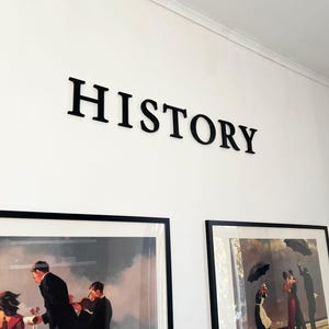 Könnte beinhalten: Schwarze Wandbuchstaben, die das Wort HISTORY in einer Serifenschrift bilden, angebracht über zwei gerahmten Kunstdrucken. Der linke Druck zeigt Figuren in formeller Kleidung, der rechte Druck zeigt Figuren mit Regenschirmen am Strand.