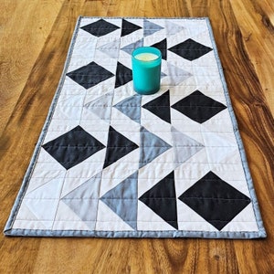 May include: A rectangular quilted table runner with a geometric pattern of black, gray, and white diamonds and triangles. A teal candle in a glass holder sits on the runner. The runner is on a wooden table.