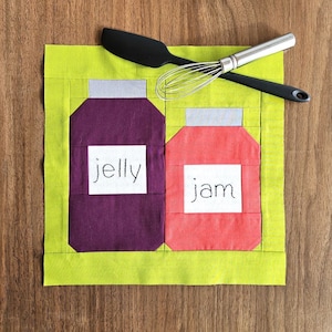 Pode incluir: Um quadrado de tecido verde com dois frascos, um com a etiqueta "jelly" e o outro com a etiqueta "jam". Uma espátula preta e um batedor de arame prateado estão em cima do tecido.