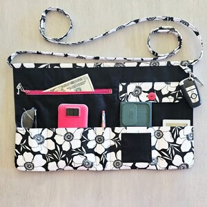 May include: Black and white floral fabric utility apron with multiple pockets. The apron has a pink phone case, a green pocket organizer, a black wallet, a key chain with a key, and a pair of glasses in the pockets.