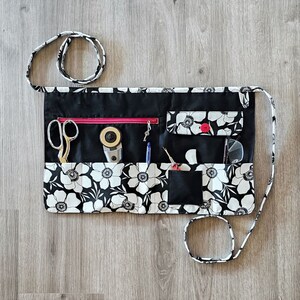 May include: A black and white floral fabric tool roll with multiple pockets and a zippered closure. The roll is open and contains various tools, including scissors, a rotary cutter, a pen, and a needle.