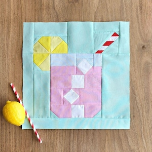 May include: A light blue fabric square with a pink and white patchwork design of a glass of lemonade with a yellow lemon wedge and a red and white striped straw.