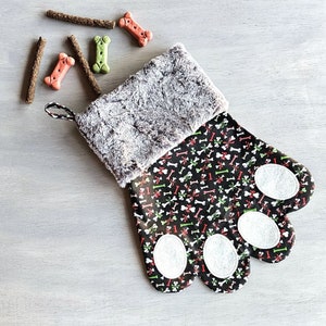 May include: A dog paw-shaped Christmas stocking with a black background and a pattern of white bones, red bows, and green accents. The cuff is gray faux fur, and white paw pads are sewn on the bottom. Dog treats are scattered around the stocking.