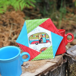 以下が含まれることがあります： 赤、緑、青、白のパッチワーク模様の六角形の布製コースター。コースターの中央には、犬が乗ったビンテージのキャンピングカーと「Happy Camper」の文字が描かれています。