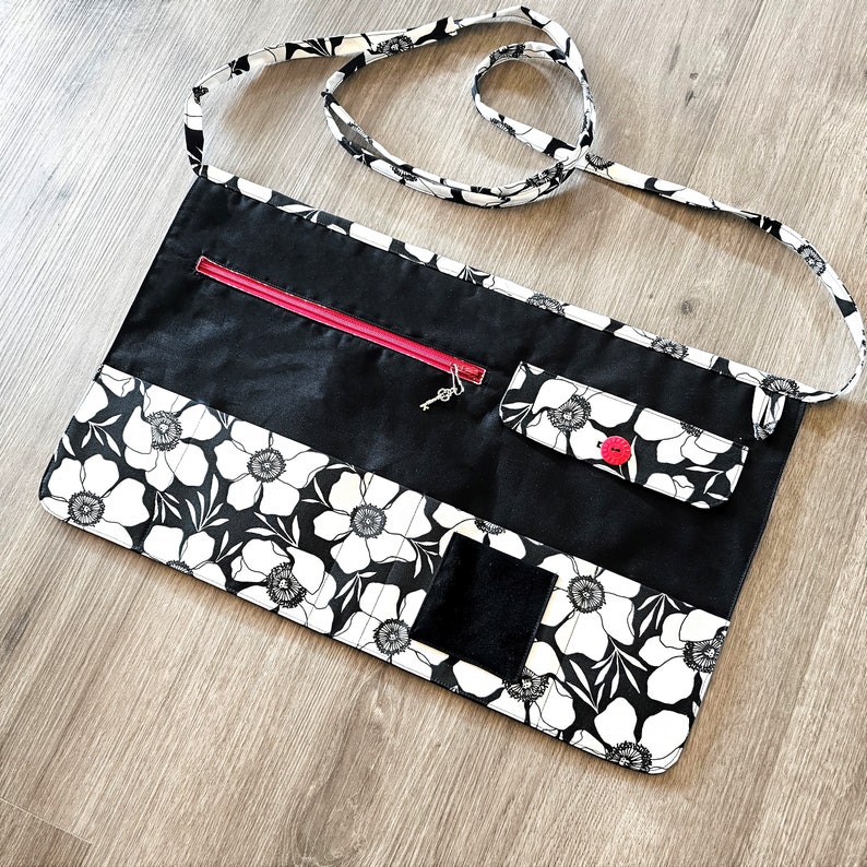 May include: Black apron with a white floral print and multiple pockets. The apron has a red zipper and a black button.